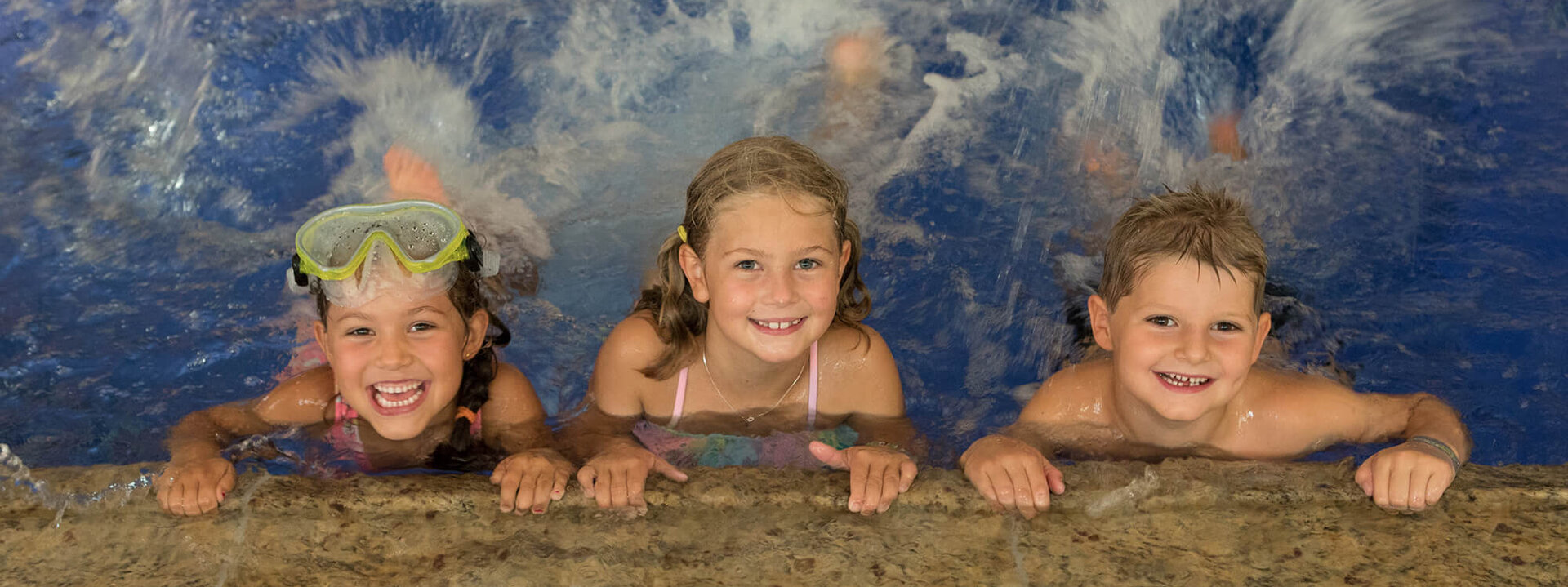 Drei fröhliche Kinder im Wasser, die am Beckenrand lächeln und mit Wasser spritzen, während sie Schwimmbrillen tragen.