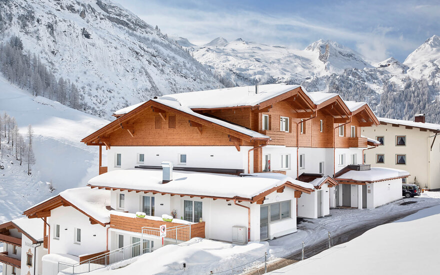 Eine moderne alpine Unterkunft mit Holzverkleidung, umgeben von schneebedeckten Bergen und klarem Himmel.
