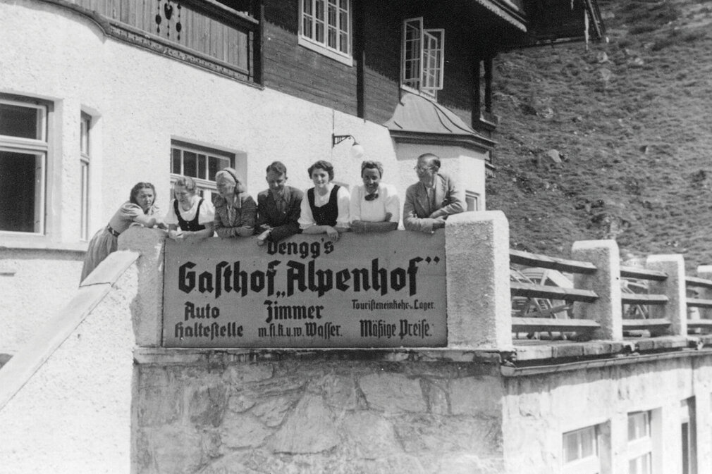 Sechs Personen lehnen an einer Mauer mit einem Schild, das den Gasthof 'Alpenhof' bewirbt, in einer bergigen Umgebung.