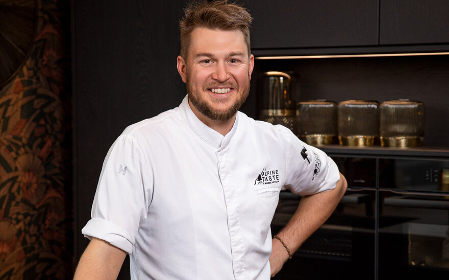 Ein lächelnder Koch in einer modernen Küche trägt eine weiße Kochjacke mit dem Logo 'Alpine Taste'.