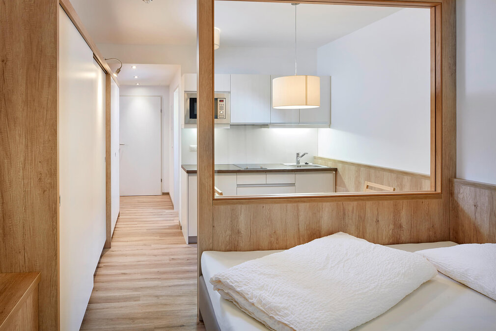 Der Raum zeigt ein gemütliches Bett mit weißen Kissen und eine offene Küche, die durch eine Holzverkleidung getrennt ist.
