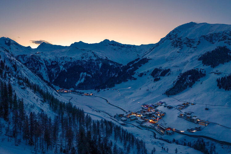 Die schneebedeckten Berge umgeben ein kleines, beleuchtetes Dorf in der Dämmerung, das eine friedliche Winteratmosphäre ausstrahlt.