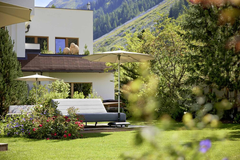 Ein elegantes Gartenmöbelstück mit einem Sonnenschirm steht in einem blühenden Garten, umgeben von grünen Bäumen und Bergen.
