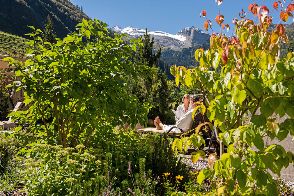 Ein Paar entspannt in einem Liegestuhl, umgeben von üppigem Grün und Blick auf schneebedeckte Berge im Hintergrund.