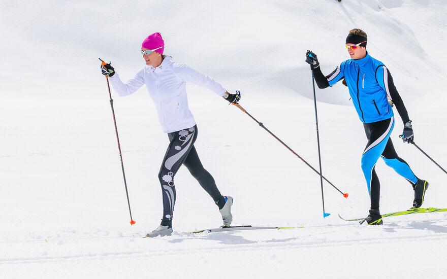 Zwei Skifahrer gleiten über eine schneebedeckte Landschaft, während die Sonne hell am Himmel scheint.