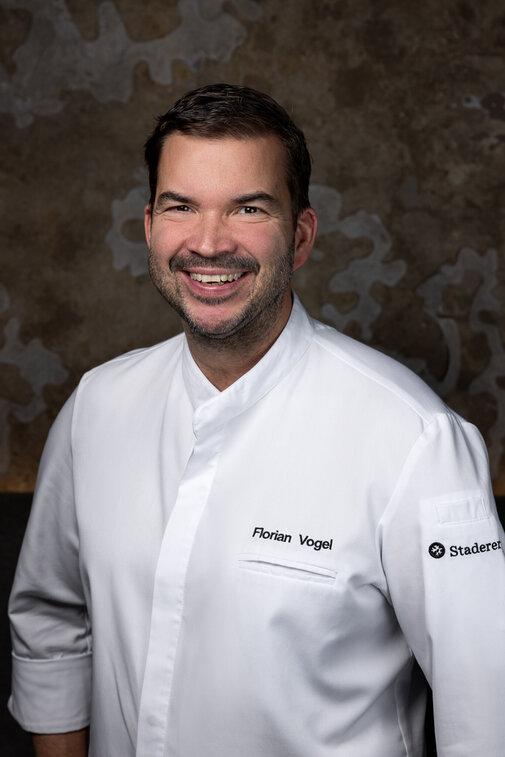 Ein lächelnder Koch trägt eine weiße Kochjacke mit seinem Namen und einem Logo auf der Brust, vor einer strukturierten Wand.