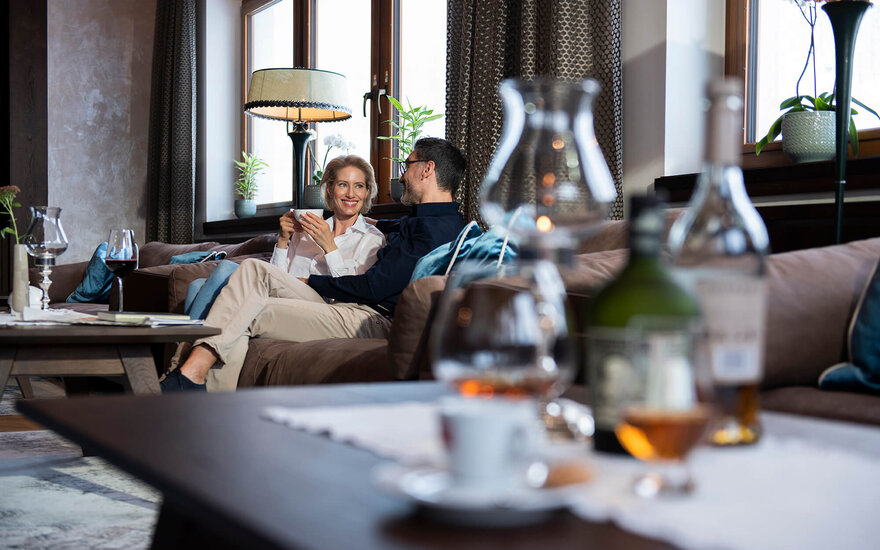 Ein Paar sitzt entspannt auf einem Sofa, während sie sich liebevoll anlächeln und eine Tasse in der Hand halten.