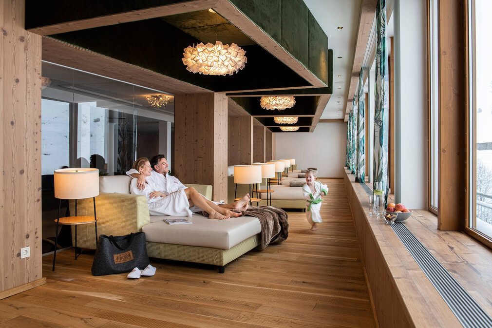 Ein Paar in Bademänteln entspannt auf einer Couch, während ein Kind fröhlich im Spa-Bereich herumläuft.