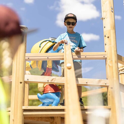 Ein Junge mit Sonnenbrille steht auf einem Spielturm und hält einen Ball in der Hand, während er lächelt.