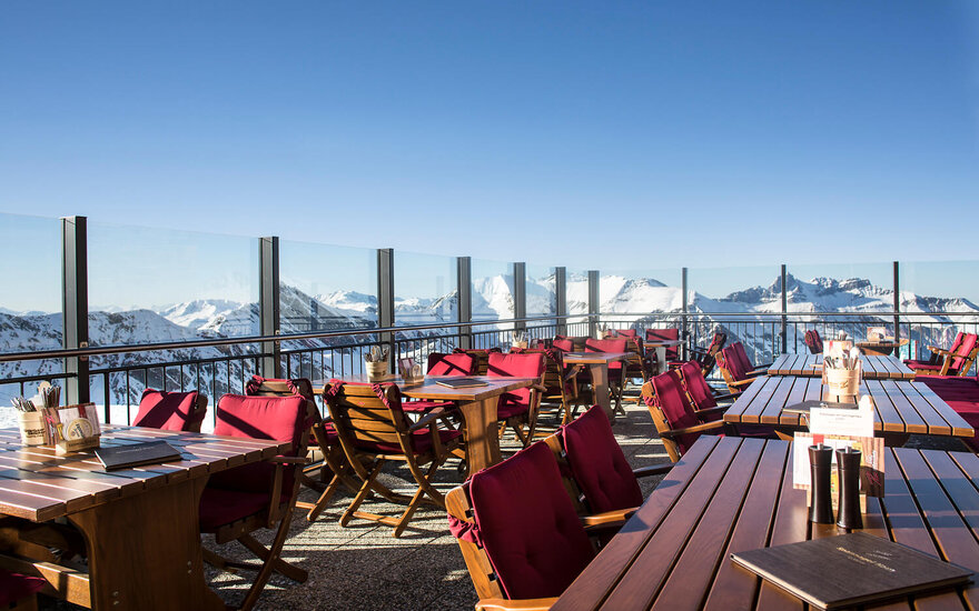Eine sonnige Terrasse mit Holztischen und roten Stühlen bietet einen atemberaubenden Blick auf schneebedeckte Berge.
