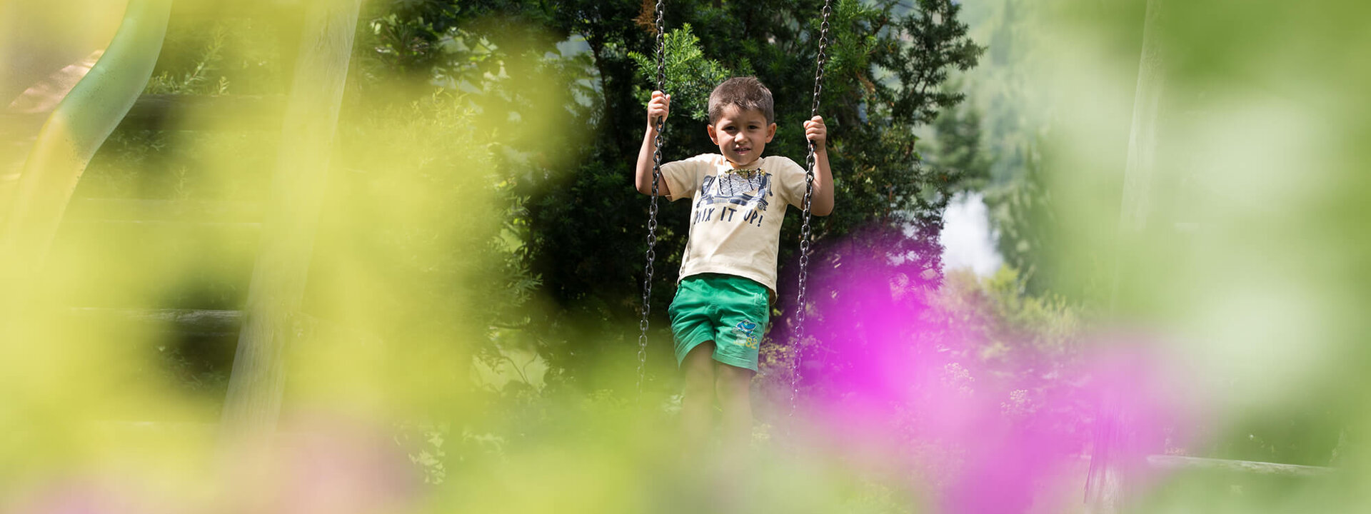 Ein fröhlicher Junge schwingt auf einer Schaukel, umgeben von bunten Blumen und grünem Laub im Hintergrund.