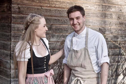 Eine junge Frau in traditioneller Tracht lächelt einen Mann in Kochkleidung an, während sie vor einer Holzwand stehen.