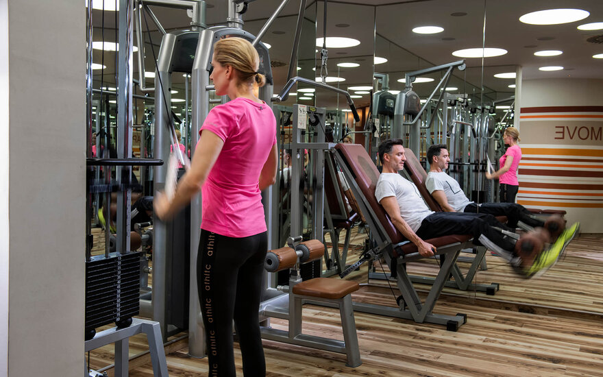 Eine Frau in einem Fitnessstudio trainiert an einem Kraftgerät, während zwei Männer auf einer anderen Maschine sitzen und sich unterhalten.