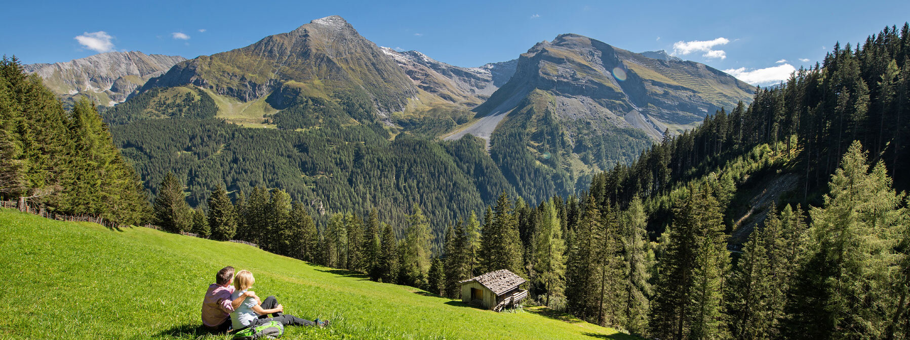 Ein Paar sitzt auf einer Wiese und genießt die atemberaubende Aussicht auf die Berge und den Wald im Hintergrund.