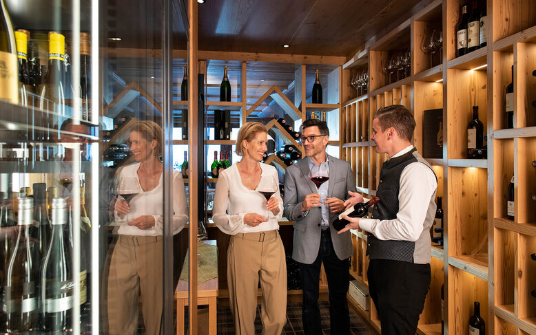 In einem eleganten Weinkeller unterhalten sich ein Paar und ein Sommelier über Weinauswahl, während sie Gläser in der Hand halten.