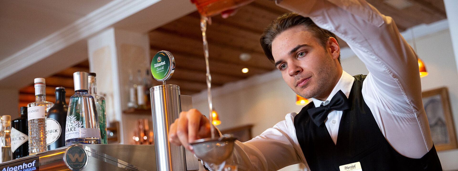 In einer stilvollen Bar bereitet ein junger Mann mit Fliege einen Cocktail vor, während er sorgfältig aus einem Shaker gießt.