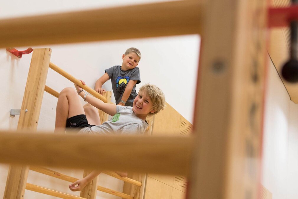 Zwei Kinder spielen fröhlich an einer Sprossenwand in einer Sporthalle, beide lächeln und haben Spaß.