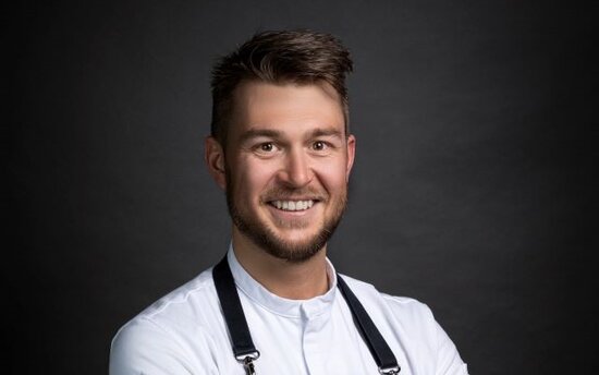 Ein lächelnder Koch in einem weißen Hemd und schwarzem Schürze steht mit verschränkten Armen vor einem dunklen Hintergrund.