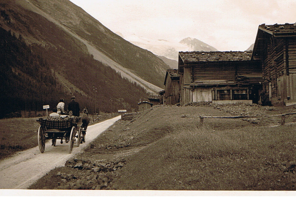 Zwei Menschen sitzen in einer Pferdekutsche, während sie durch eine malerische Berglandschaft mit alten Gebäuden fahren.