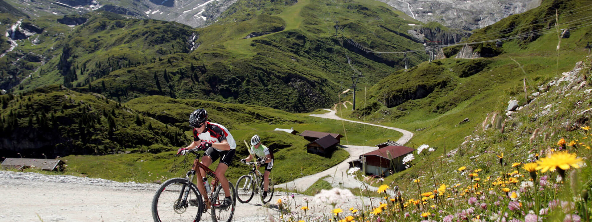 Zwei Mountainbiker erklimmen einen steilen Weg in den Zillertaler Alpen, umgeben von blühenden Wiesen und schneebedeckten Gipfeln.