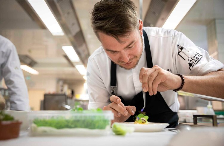 Ein Koch verziert sorgfältig einen Teller mit frischen Kräutern und essbaren Blumen in einer modernen Küche.