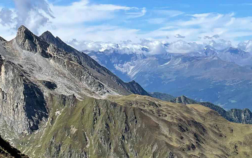 Majestätische Berglandschaft mit schroffen Gipfeln und sanften Hügeln, unter einem strahlend blauen Himmel mit Wolken.