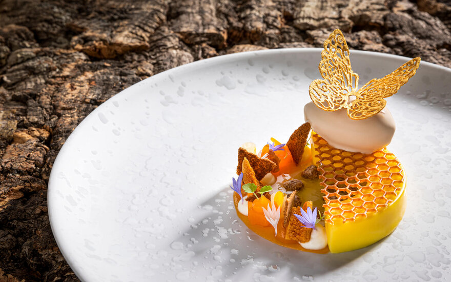 Ein kunstvoll angerichtetes Dessert mit einer goldenen Schmetterlingsdekoration und bunten essbaren Blüten auf einem weißen Teller.