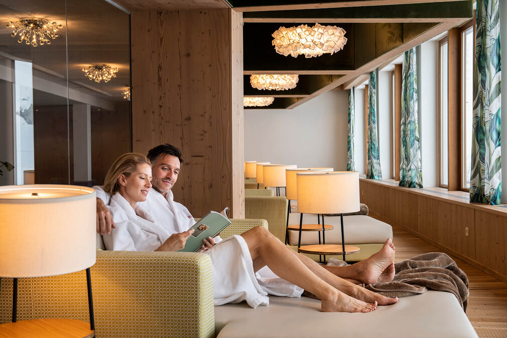 Ein Paar in Bademänteln entspannt auf einer Couch und liest gemeinsam in einem Buch, umgeben von warmem Licht.
