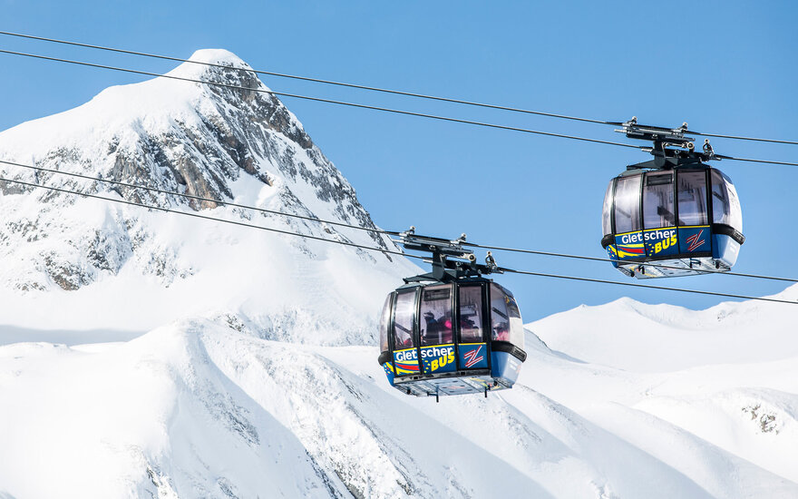 Zwei moderne Gondeln schweben über schneebedeckte Berge, während der klare Himmel im Hintergrund strahlt.
