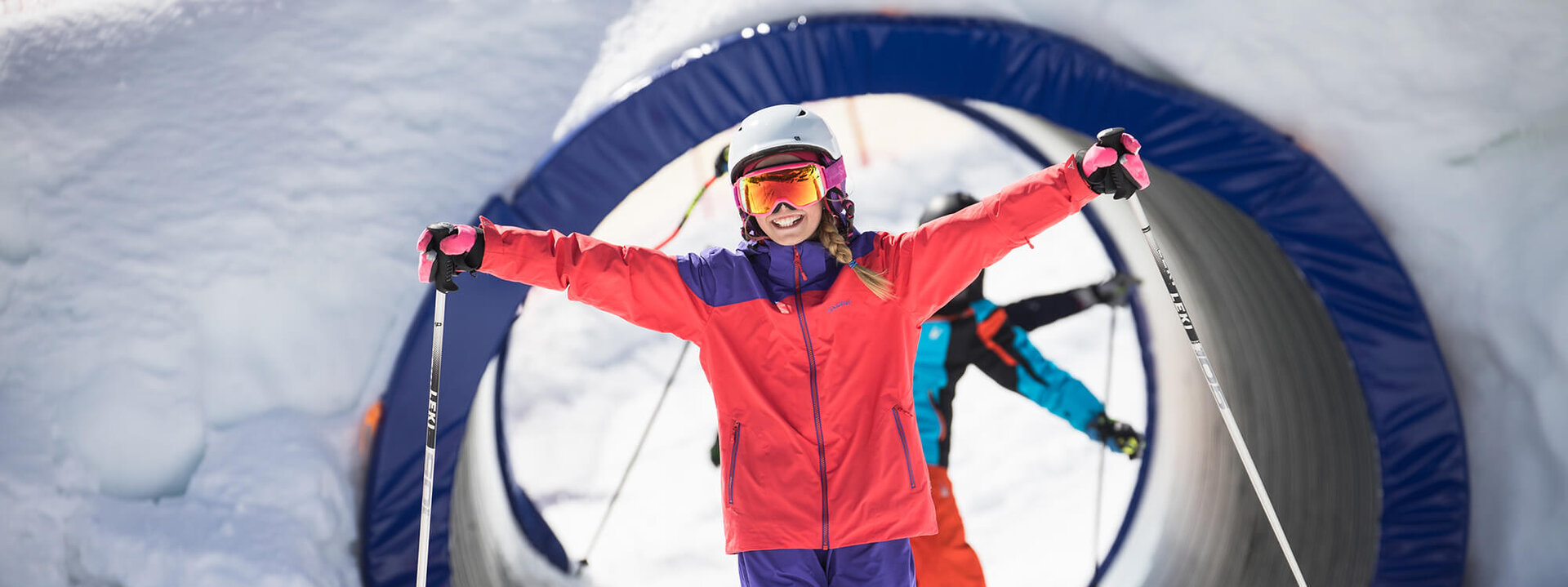 Eine fröhliche Skifahrerin in leuchtender Kleidung posiert mit ausgebreiteten Armen auf einer schneebedeckten Piste.