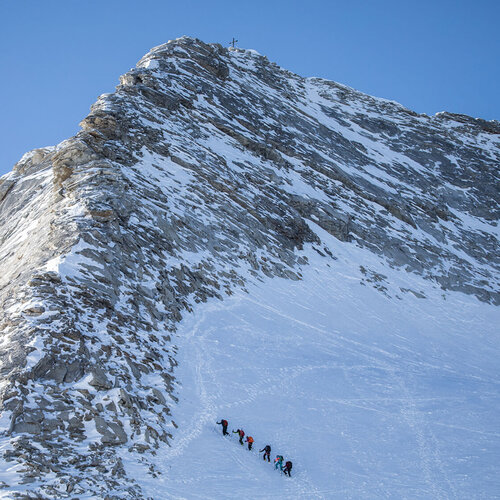 Eine Gruppe von Bergsteigern erklimmt einen schneebedeckten Gipfel unter einem strahlend blauen Himmel.