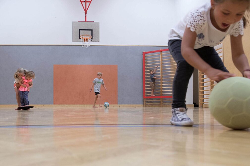 In einer Sporthalle spielen Kinder, während eines ein Mädchen auf einem Spielgerät sitzt und ein anderes den Ball rollt.