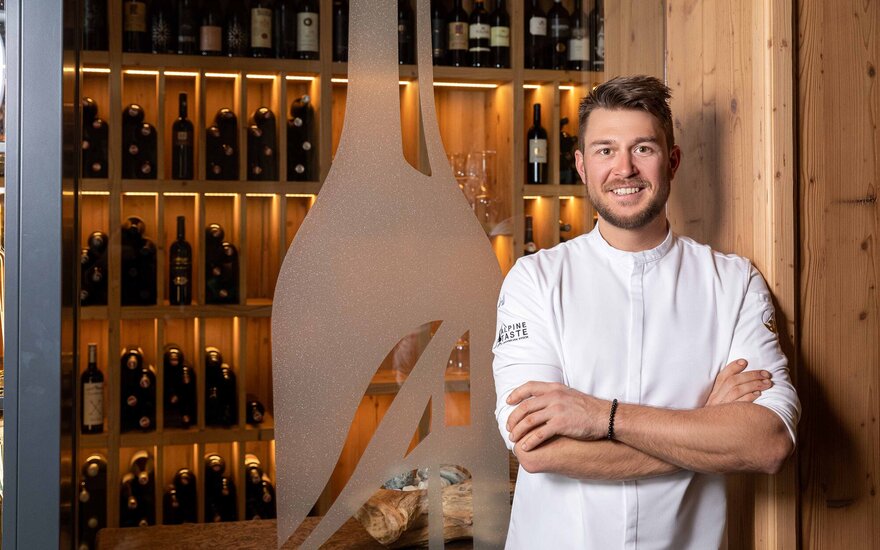 Ein lächelnder Koch in einem weißen Outfit steht vor einer Wand mit beleuchteten Weinflaschen in einem rustikalen Ambiente.