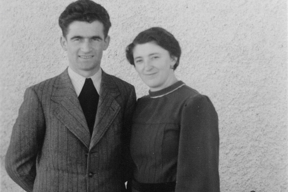 Das Bild zeigt ein Paar in formeller Kleidung, der Mann mit Anzug und Krawatte, die Frau in einem langen, eleganten Kleid.