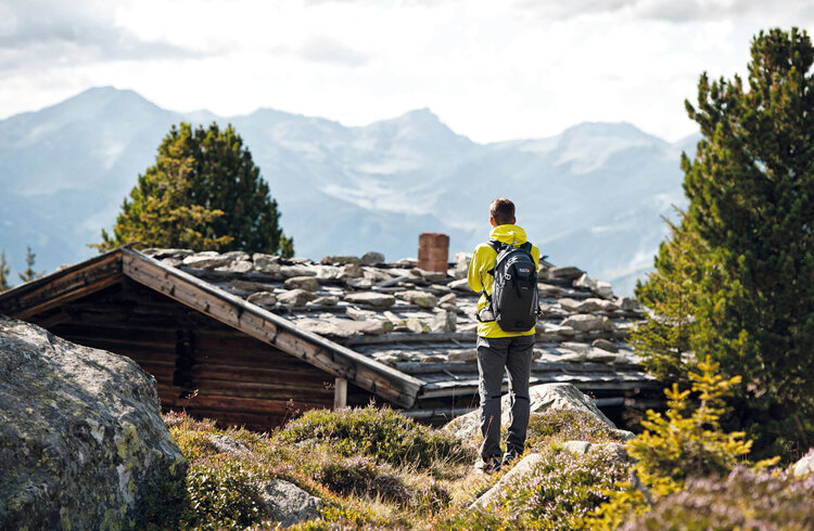 Ein Wanderer in gelber Jacke steht vor einer Berghütte und blickt auf die majestätischen Alpen im Hintergrund.