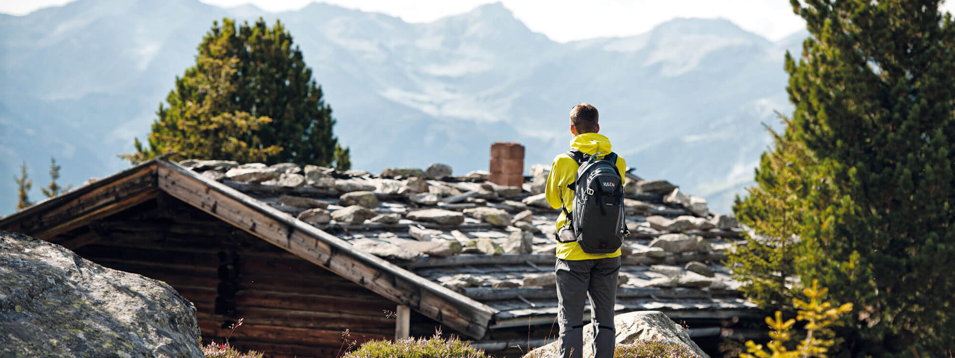 Ein Wanderer in gelber Jacke steht vor einer Berghütte und blickt auf die majestätischen Alpen im Hintergrund.